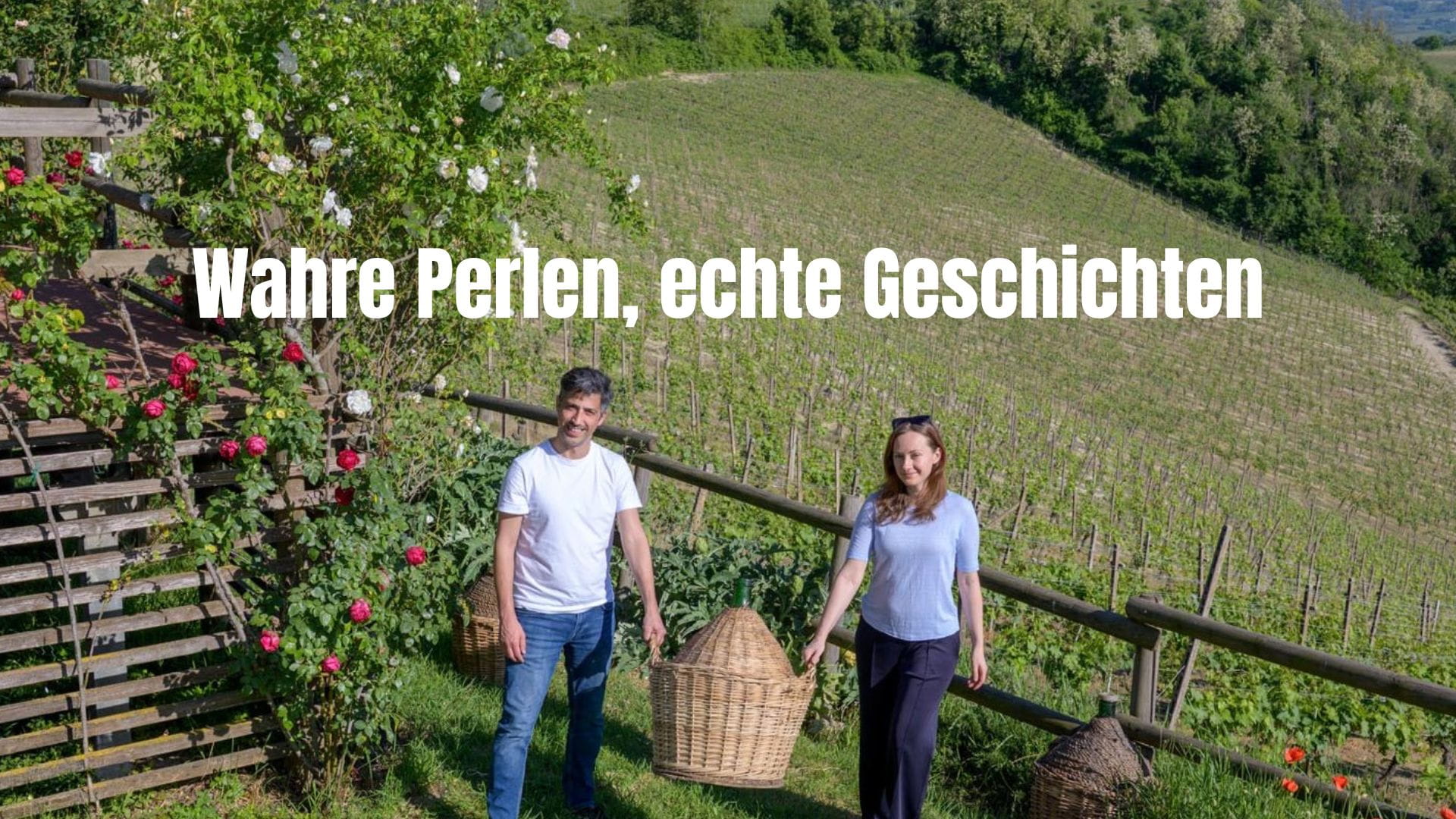 Ein Mann und eine Frau stehen lächelnd mit einem großen geflochtenen Weinkorb (Demijohn) vor einem Weinberg, im Hintergrund sanfte Hügel und ein Rosenstrauch.