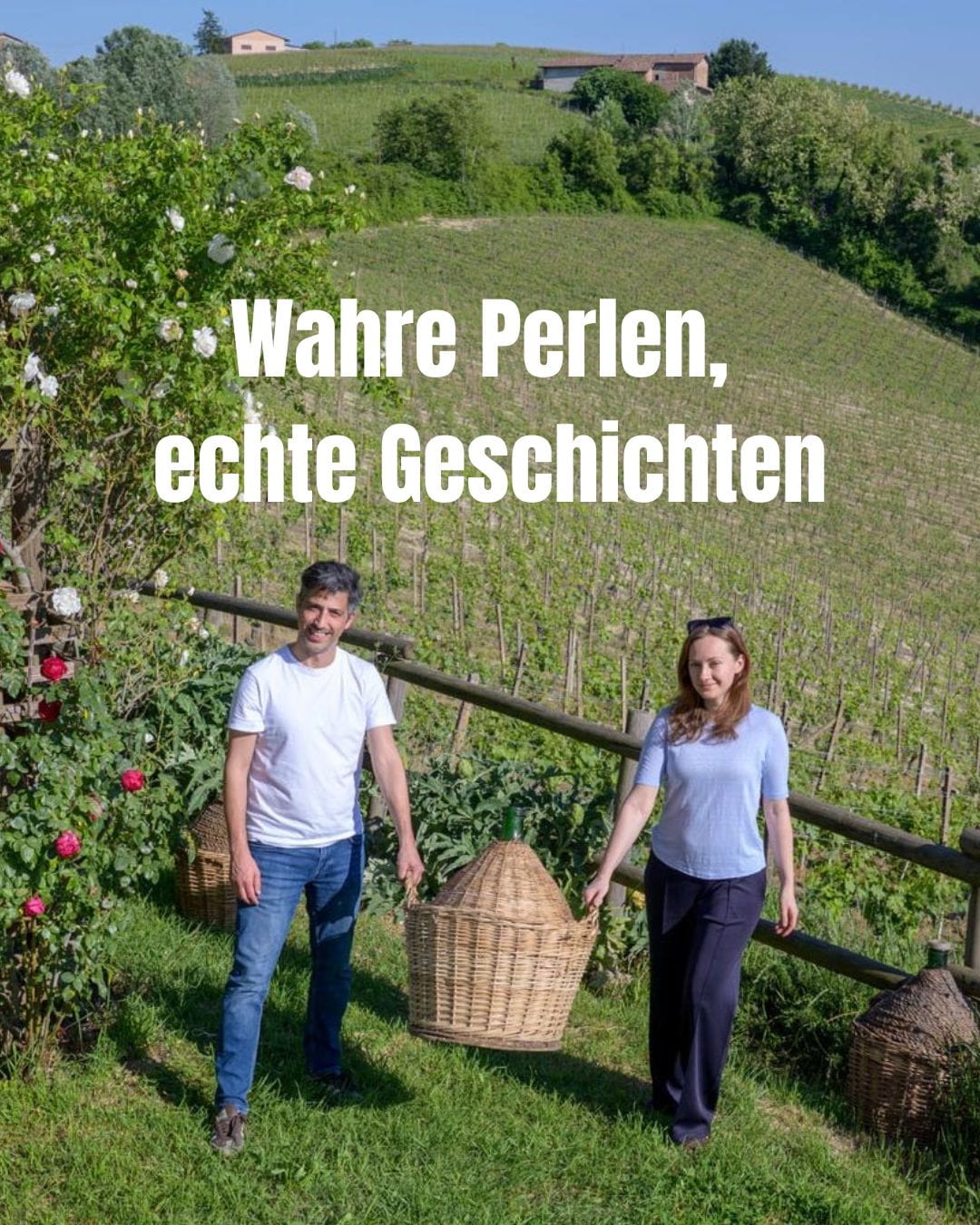 Ein Mann und eine Frau stehen lächelnd mit einem großen geflochtenen Weinkorb (Demijohn) vor einem Weinberg, im Hintergrund sanfte Hügel und ein Rosenstrauch.
