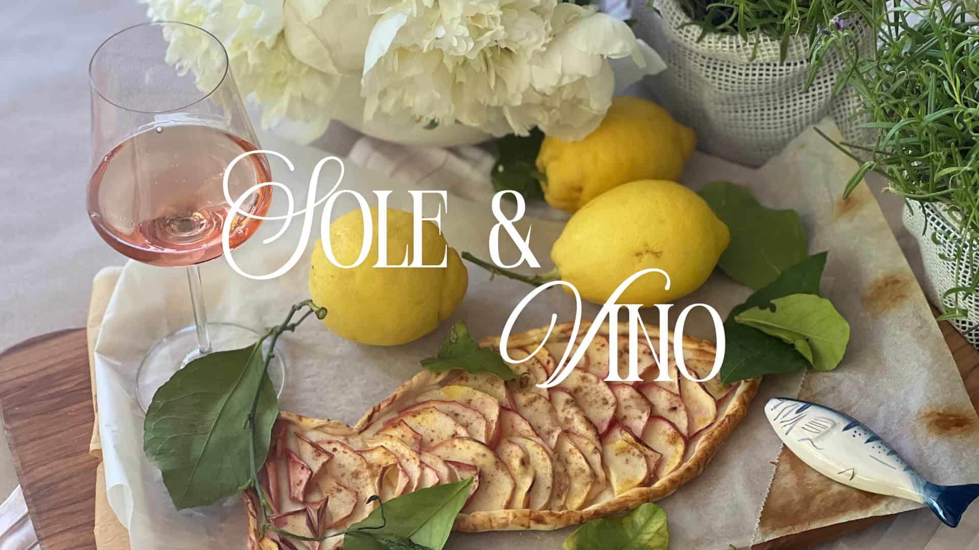 Italienische Weine und Delikatessen - Ein sommerlich gedeckter Tisch mit einem Glas Roséwein, frischen Zitronen, einer apfelgefüllten Blätterteig-Tarte in Fischform und weißen Pfingstrosen im Hintergrund. Über dem Bild liegt der geschwungene Schriftzug „SOLE & VINO“